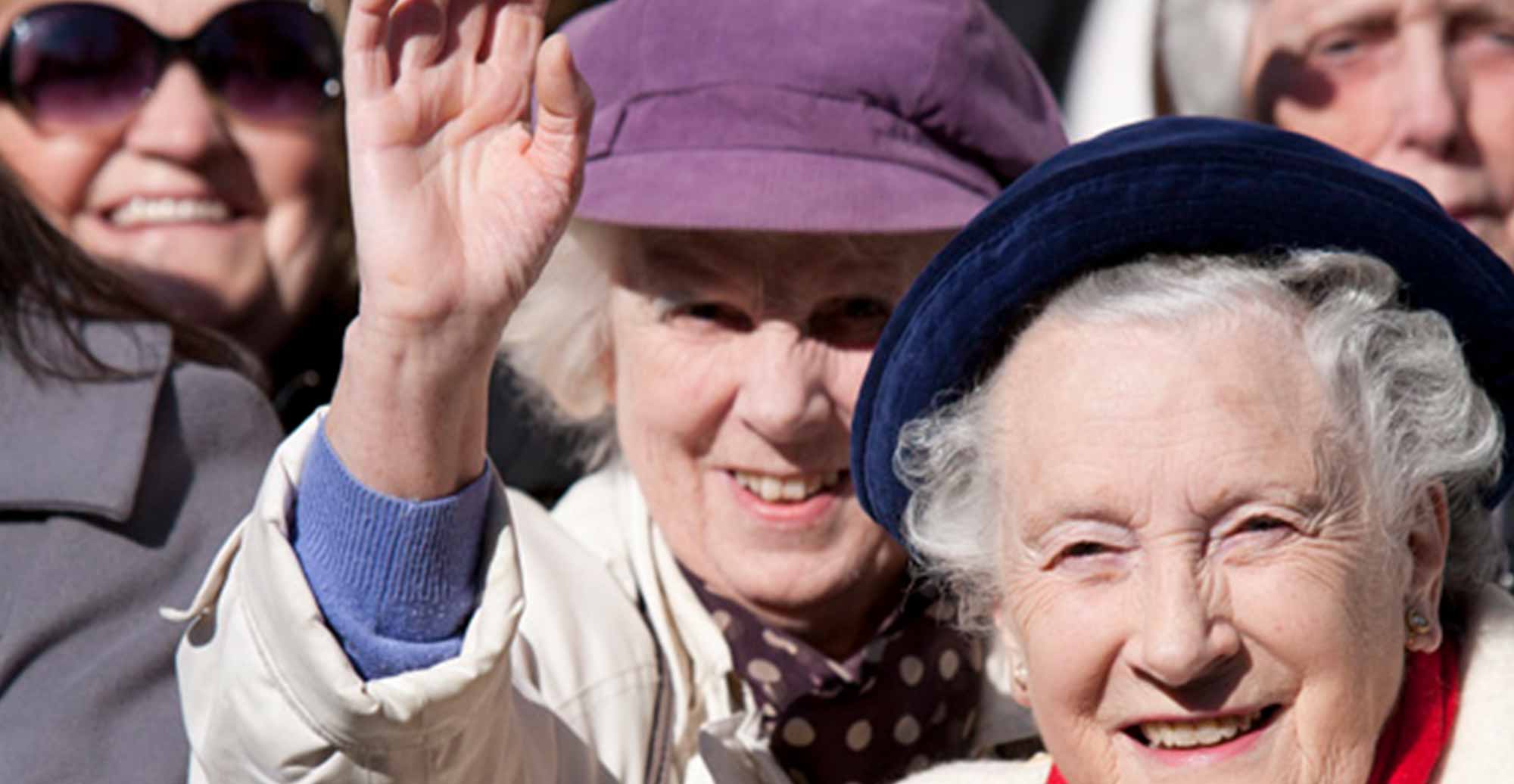 A crowd of people smiling but the focus is on a lady in a purple hat who waves to the camera.