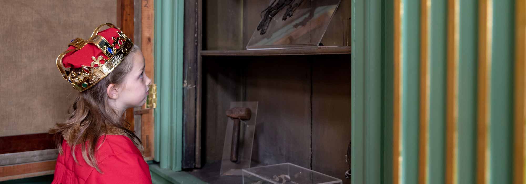 A little girl dressed in a crown and cloak gazes at a display of objects in the Mansion House.