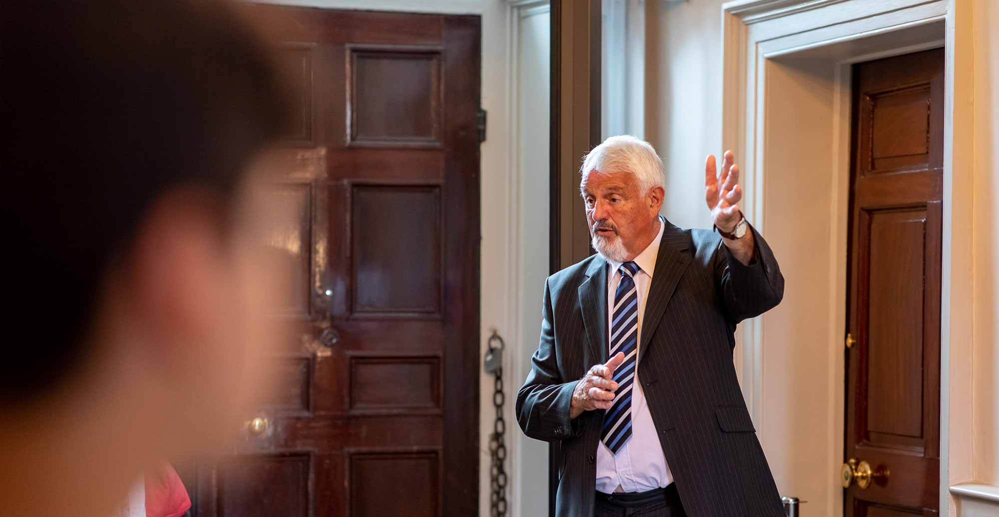 A man in a suit talks to a group of visitors at the Mansion House.