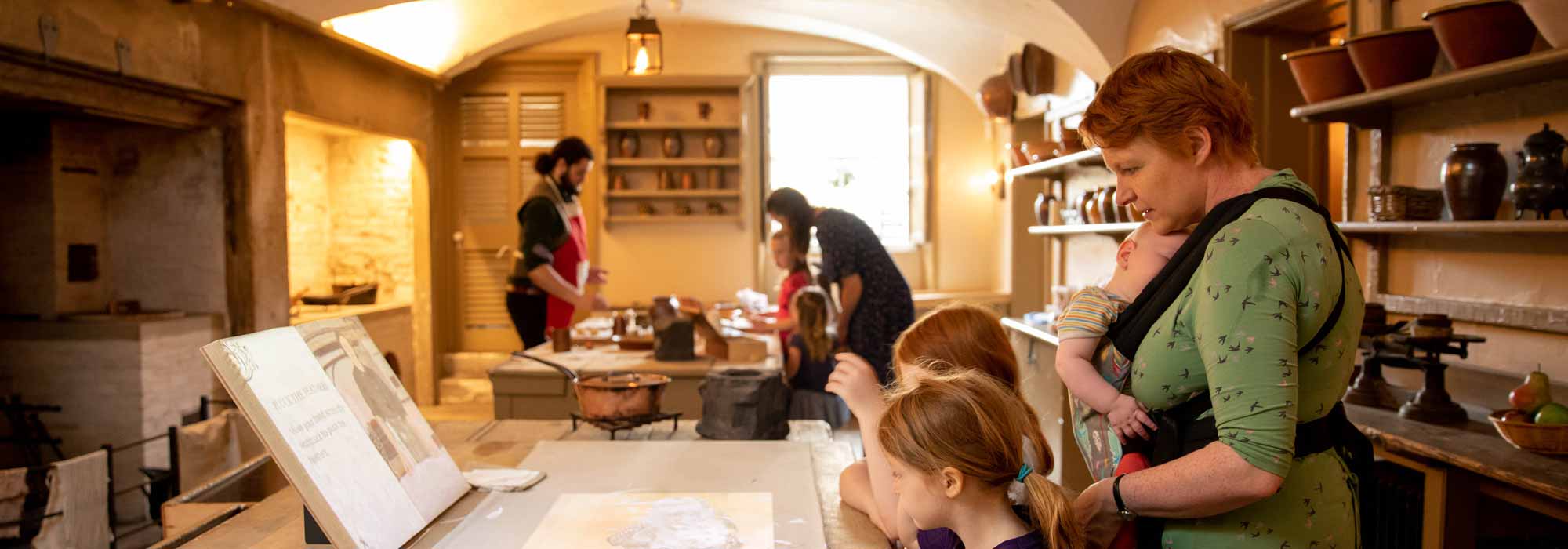 A lady with a baby in a baby carrier oversees some children learning how to bake a recipe by following a cookery book.