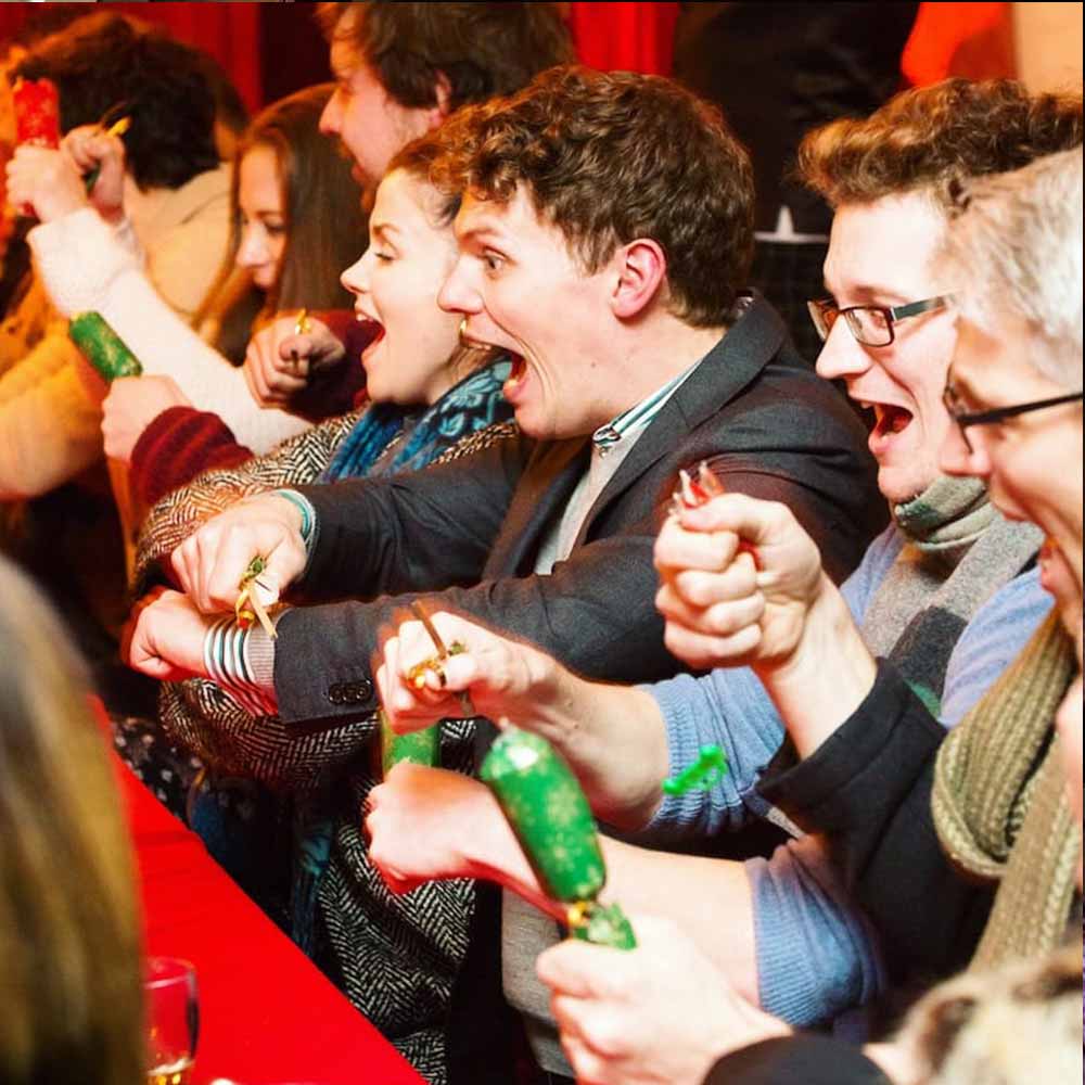 A row of people excitedly cheer whilst popping Christmas crackers.
