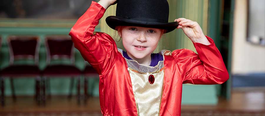 A little girl with a red and white costume dress places a bowler hat on her head whilst smiling