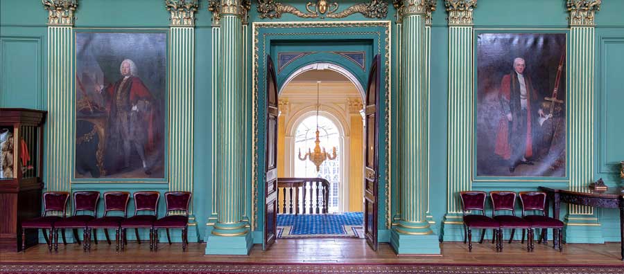 A view of the doorway of one of the rooms in the Mansion House which leads to a stairway. The room is green and on either side of the door way hangs a large portrait and beneath these some chairs have been placed with their backs against the wall