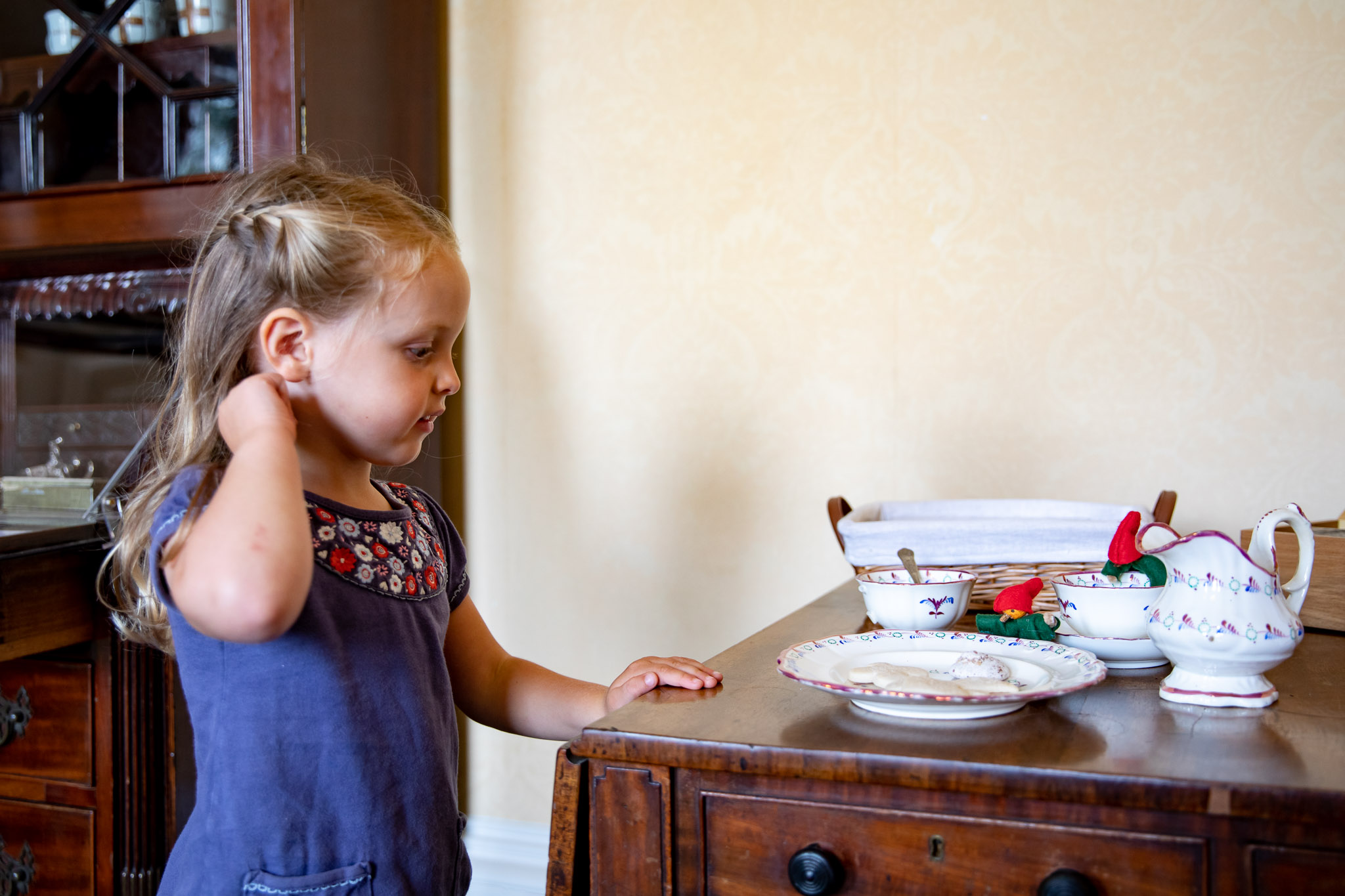 Little girl in blue dress looks at tea set on a dark wood table
