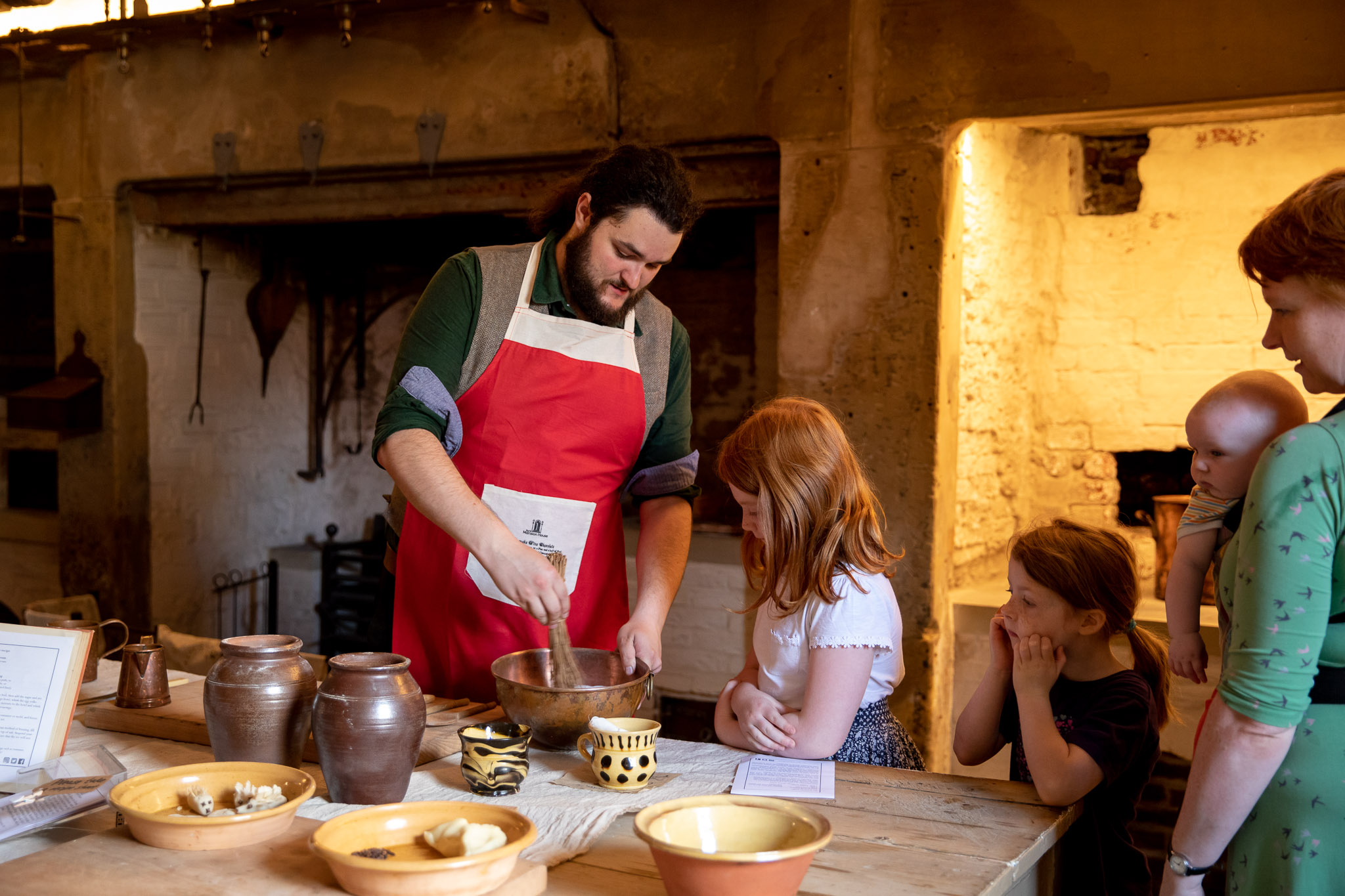 A man is demonstrating how to cook to two young children and a lady with a baby in a carrier