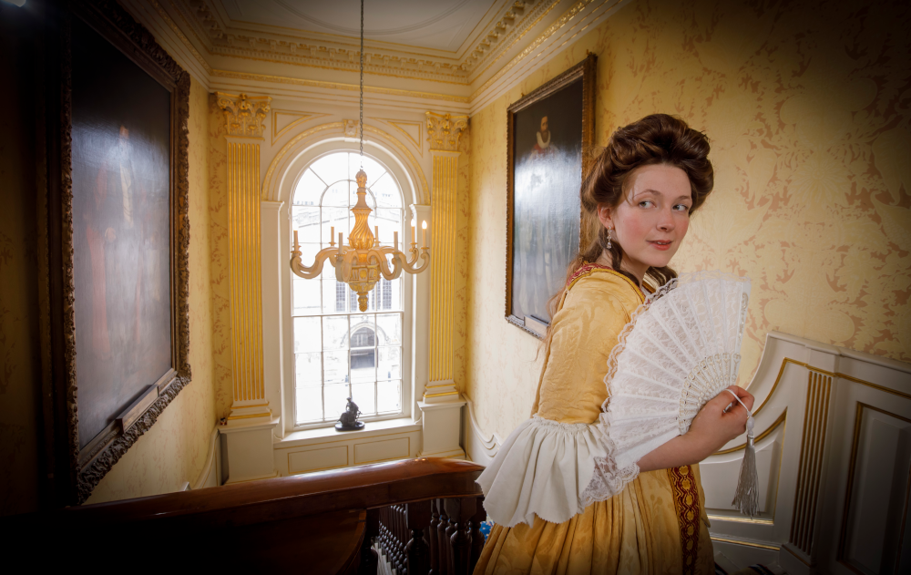 A lady in a Georgian-era yellow dress holds a fan