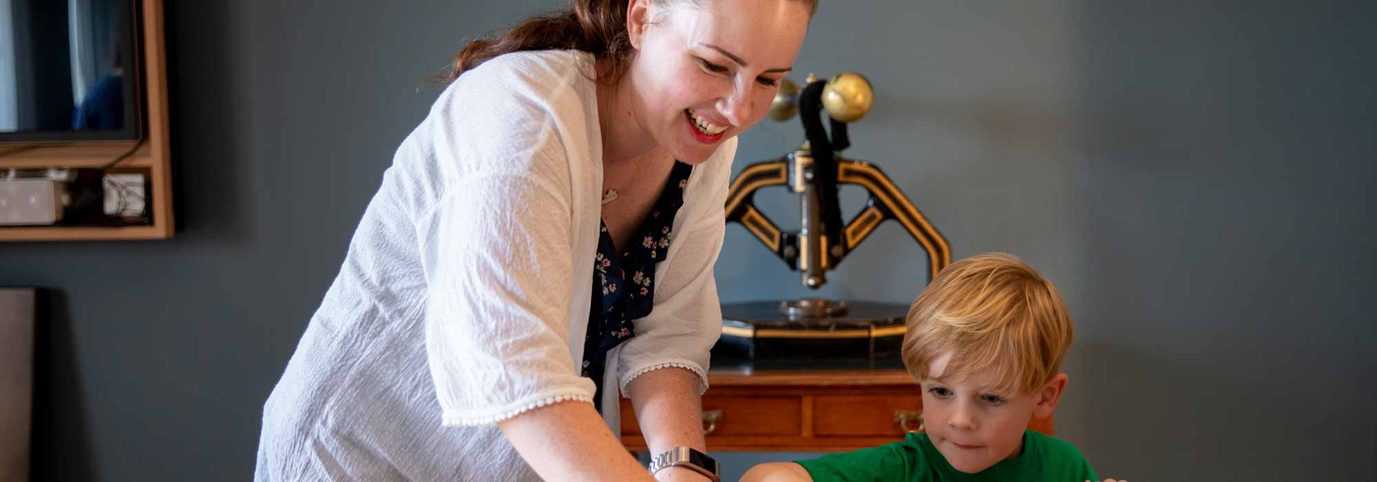 A woman helping a young boy at a table to complete a task. Both are smiling.