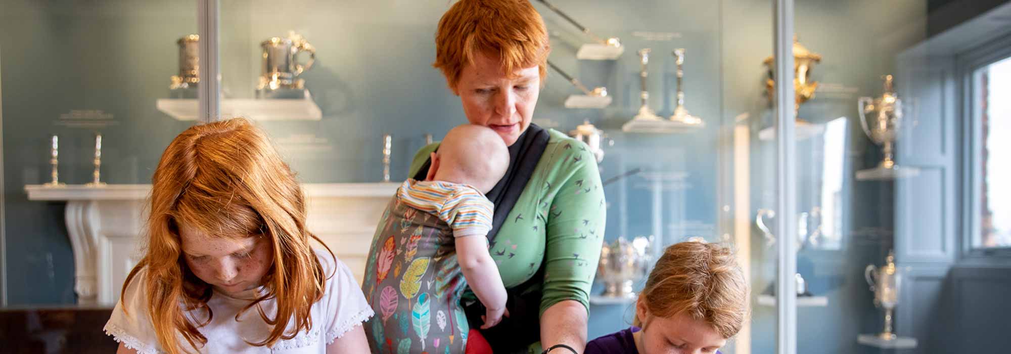 A lady with a baby in a baby carrier helps two young children to complete a task at a table.