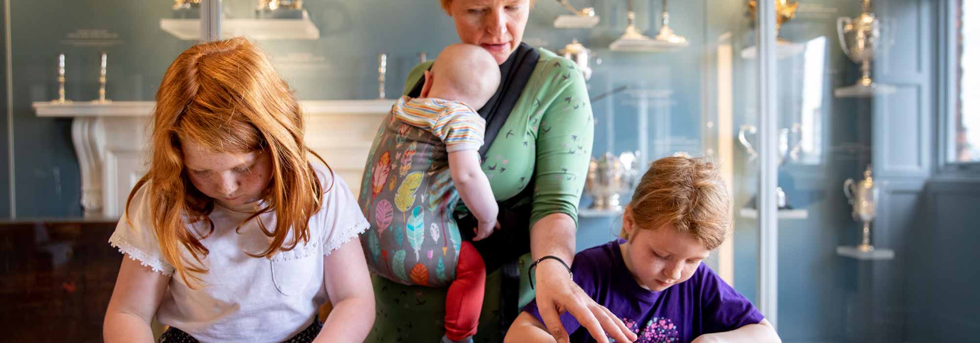 Two young children complete a task whilst a woman in a green shirt with a baby in a baby carrier supervises them